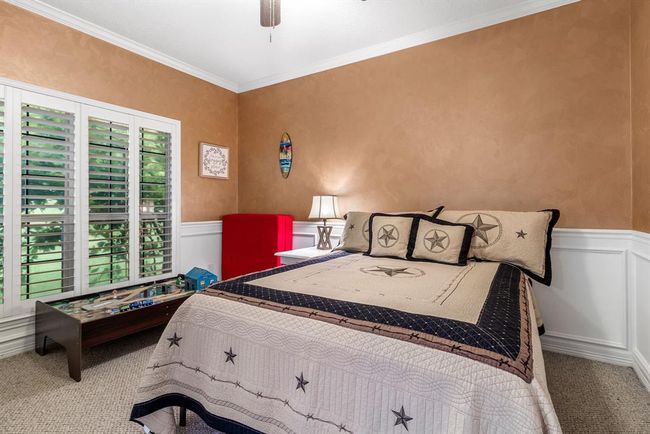 This cozy bedroom features warm tan walls and white wainscoting, with plantation shutters on the window. It includes a comfortable bed with star-themed bedding, a side table with a lamp, and a red cabinet. A small table with a train set adds a playful touch. | Image 4