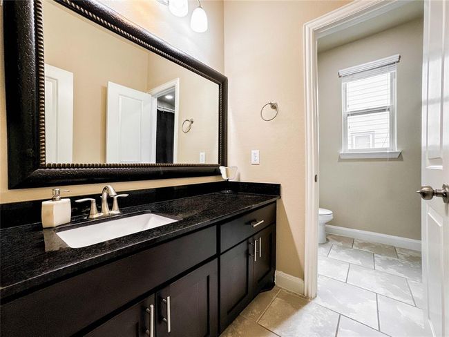 This bathroom features a sleek dark vanity with a large mirror, modern fixtures, and ample storage. The tiled floor leads to a separate toilet area with natural light from a window. | Image 38