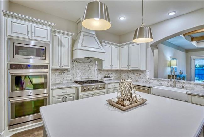 This modern kitchen features elegant white cabinetry, stainless steel appliances, and a spacious island with stylish pendant lighting. The open layout flows into the adjacent living area, creating a welcoming and functional space for cooking and entertaining. | Image 26