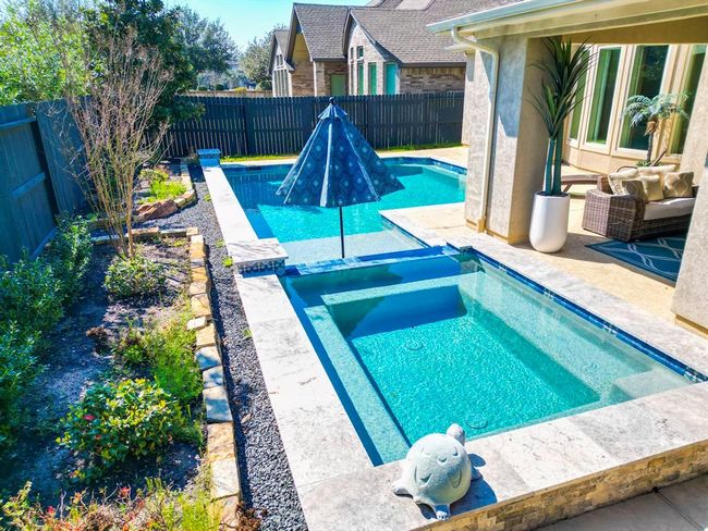 This photo showcases a modern backyard featuring a sparkling pool and spa area. The pool is adjacent to a landscaped garden with a stone border. A covered patio with comfortable seating offers a relaxing outdoor space, perfect for entertaining. | Image 4
