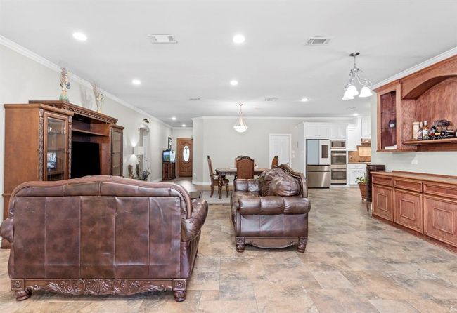 This is a spacious, open-plan living area featuring recessed lighting, tiled flooring, and a neutral color palette. The room is well-appointed with a large entertainment unit, comfortable leather seating, and transitions seamlessly into a dining space and a kitchen with wooden cabinetry and stainless steel appliances. | Image 19