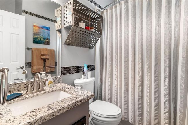 This bathroom features a modern design with a granite countertop, sleek fixtures, and a striped shower curtain. It includes a wall-mounted storage unit and tasteful decor, creating a functional and stylish space. | Image 23