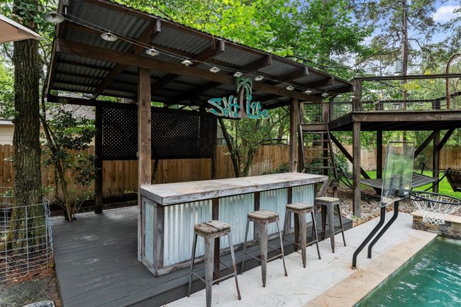 This backyard features a rustic outdoor bar with a corrugated metal counter, shaded by a wooden pergola. The area includes a poolside with a removable basketball hoop and lush greenery, perfect for entertaining and relaxation. | Image 40