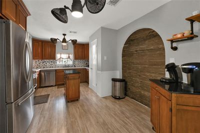 The beautifully updated kitchen features stainless steel appliances, wood cabinetry, a tiled backsplash, and wood like flooring. It also has a unique arched wooden accent wall, modern lighting fixtures, and a small island. | Image 2