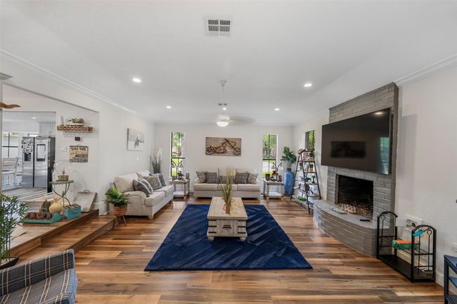 A spacious, well-lit living room with modern hardwood flooring and recessed lighting. The room includes a stylish gray brick fireplace. Large windows offer natural light, while the open layout connects to a glimpse of the dining area. | Image 4