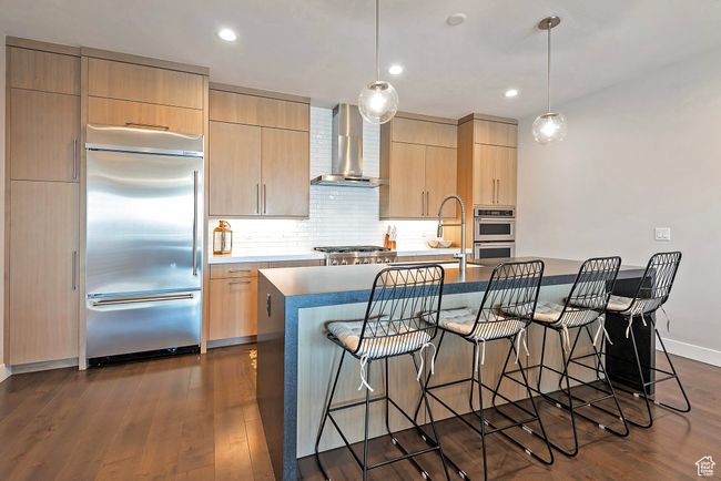 Kitchen featuring a kitchen breakfast bar, wall chimney range hood, light brown cabinets, a center island with sink, and appliances with stainless steel finishes | Image 11