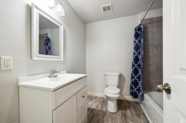 This bathroom features a modern design with a white vanity and sink, a large mirror, and a combination shower-tub with a stylish blue geometric shower curtain. The flooring is wood-style, adding a warm touch. | Image 13