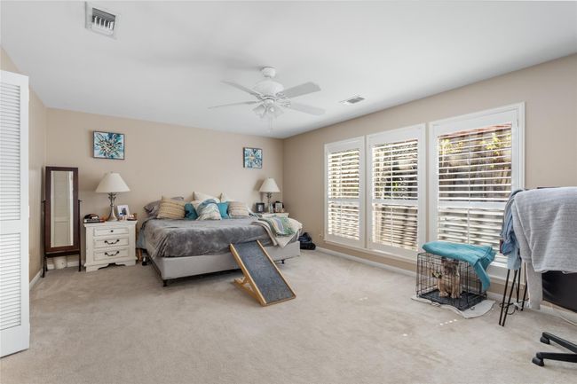 This spacious primary bedroom offers a serene retreat with soft neutral tones, plush carpeting, and abundant natural light streaming through large windows adorned with plantation shutters. The generous layout provides ample space for relaxation, while the ceiling fan ensures year-round comfort. | Image 23