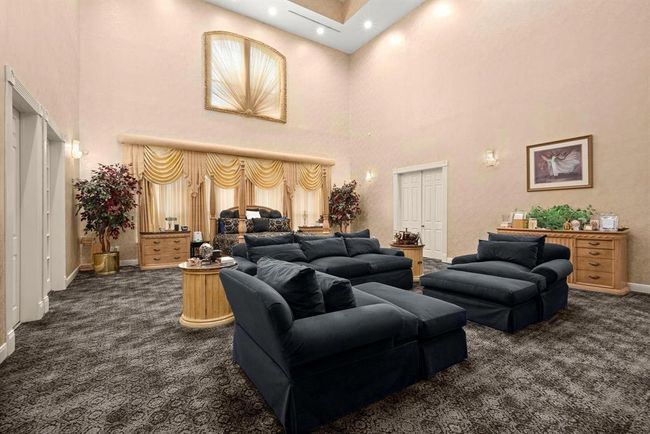This is a spacious, primary bedroom featuring plush dark sofas, elegant golden drapes, and a mixture of classic wood furniture. The room is well-lit, with both natural light from the window and recessed ceiling lights, and it boasts a high 25 ft ceiling that enhances the sense of space. It's finished with patterned carpeting for added texture and comfort. | Image 12