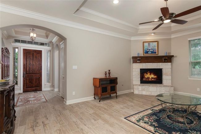 This inviting living area features a cozy stone fireplace, a ceiling fan, and elegant crown molding. The wood-like tile flooring complements the warm, neutral walls, and the entrance is accentuated by a rich wooden door and stylish archways. | Image 9