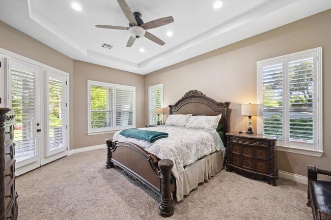 The master bedroom features a high ceiling with a fan, elegant wood furniture, and plenty of natural light through large windows with shutters. French doors lead to a serene outdoor view. | Image 9