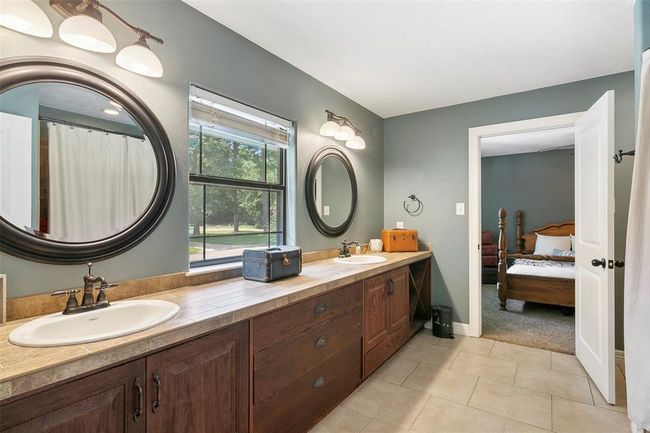 This bathroom features a spacious dual-sink vanity with dark wood cabinetry, elegant round mirrors, and modern lighting. A window provides natural light, and the room leads into a cozy bedroom. | Image 38