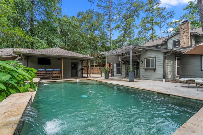 This photo showcases a serene backyard oasis featuring a spacious swimming pool with a waterfall feature, a covered outdoor lounge area, and a charming pergola attached to a modern house. Surrounded by lush greenery, it's perfect for relaxation and entertaining. | Image 38