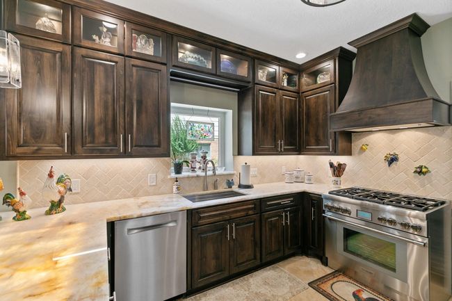 This beautifully updated kitchen showcases rich custom wood cabinetry with illuminated glass upper cabinets, quartzite countertops, and premium stainless steel appliances including a Thermador gas range, complemented by a classic herringbone tile backsplash and warm under-cabinet lighting that creates an inviting culinary workspace. | Image 8