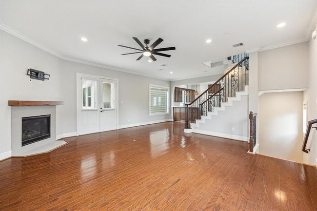 This spacious living area features hardwood floors, a cozy corner fireplace, and a modern ceiling fan. The room is well-lit with recessed lighting and has a stylish open staircase with wrought iron railing. French doors provide stylish natural light. | Image 4