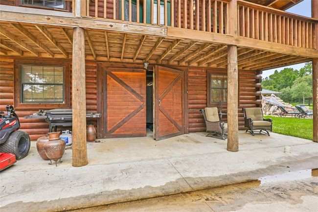 This photo showcases a rustic log cabin exterior with a covered patio, featuring sturdy wooden beams and barn doors. The area includes outdoor seating, a grill, and a view of a manicured lawn and pool, perfect for relaxation and entertaining. | Image 45