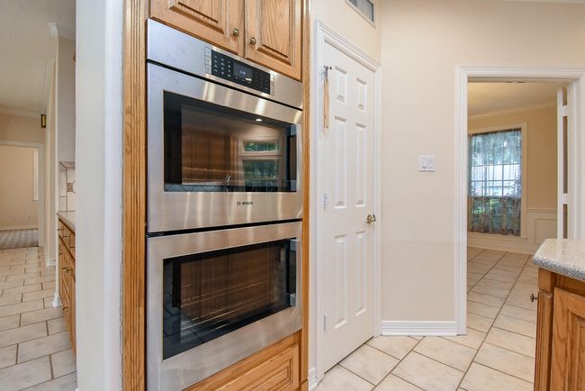 This kitchen features a double built-in oven with a stainless steel finish, surrounded by wooden cabinetry. The space is bright, with light-colored walls and tile flooring, offering a view into an adjacent room with a window. | Image 17