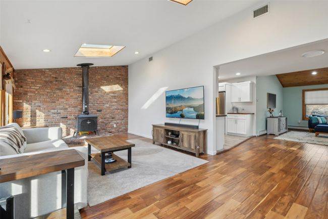 Living area with visible vents, vaulted ceiling with skylight, a wood stove, a baseboard heating unit, and light wood-type flooring | Image 4