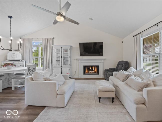 living area with a warm lit gas log fireplace, wood finished floors, vaulted ceiling, and ceiling fan with chandelier in dining area | Image 5