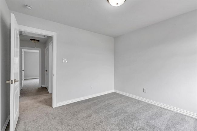 This photo shows a bright, empty room with light gray walls and carpet, featuring a simple ceiling light. The open door leads to a hallway with additional doors, suggesting access to more rooms. | Image 35