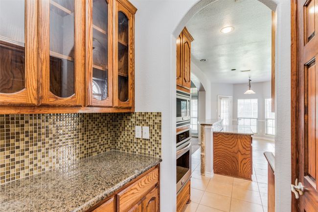 Kitchen with decorative backsplash, glass insert cabinets, light stone counters, brown cabinets, and a textured ceiling | Image 15
