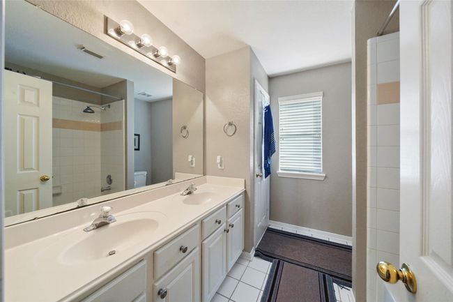 This bathroom features a double vanity with ample storage, a large mirror, and bright lighting. It includes a shower-tub combo, neutral tile flooring, and a window for natural light. | Image 26