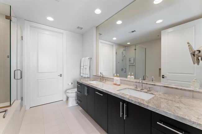 Modern bathroom with dual sinks, a large mirror, and a spacious glass shower. Features sleek dark cabinetry, elegant countertops, and bright recessed lighting for a clean and contemporary feel. Door leads to the ensuite closet. | Image 14