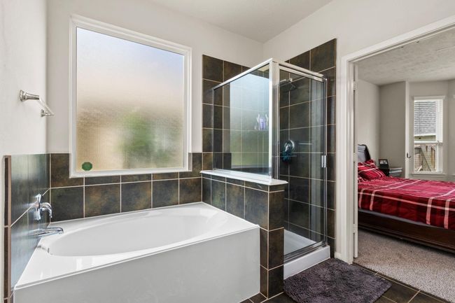 This bathroom features a large soaking tub and a separate glass-enclosed shower with dark tile accents. A frosted window provides natural light while maintaining privacy. The space connects to a bedroom with carpeted flooring. | Image 18