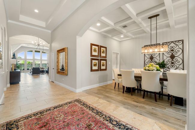 Step into elegance with this stunning entryway and formal dining room. The grand foyer welcomes you with tray ceilings, arched openings and warm tile flooring that leads to a spacious open-concept living area. To the right, a beautifully appointed dining room features a coffered ceiling and designer chandelier. With natural light streaming in and thoughtful architectural details throughout. | Image 9