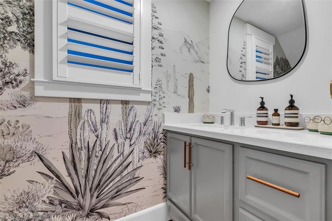 This powder room features a desert-themed wallpaper with cacti, a sleek gray vanity with modern hardware, and a stylish oval mirror. The white countertop and window shutters add brightness to the space. | Image 19