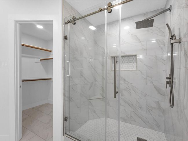 This photo showcases a modern bathroom with a spacious walk-in shower featuring elegant marble-style tiles, a rainfall showerhead, and sliding glass doors. Adjacent is a well-lit walk-in closet with ample shelving and hanging space. | Image 25