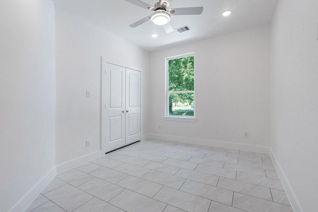 This room features bright white walls and tiled flooring, creating a fresh and modern feel. It includes a large window for natural light, a ceiling fan, and a closet with double doors, perfect for additional storage. | Image 27