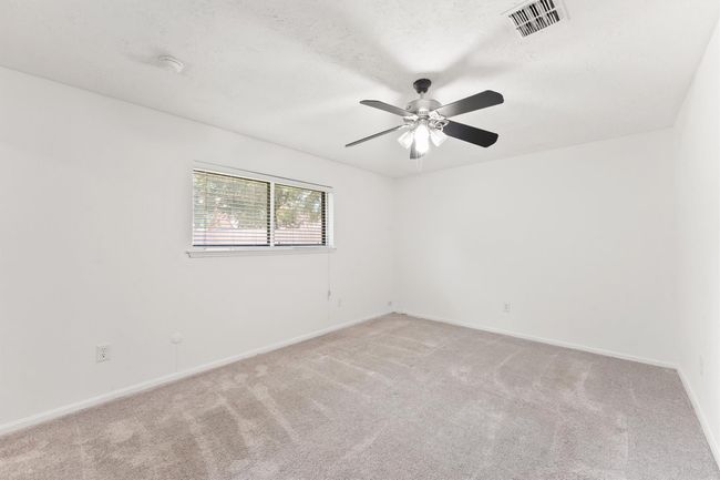 This is a bright, spacious bedroom featuring fresh white walls, plush carpet, a ceiling fan with lights, and a large window for natural light. | Image 19