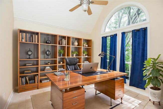 Spacious room with tiled floors and beige walls, featuring large arched windows with blue curtains. Abundant natural light and a ceiling fan enhance the airy feel. *Virtually staged | Image 12