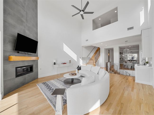 Living area with a towering ceiling, a tiled fireplace, a chandelier, a ceiling fan, and light wood-type flooring | Image 16