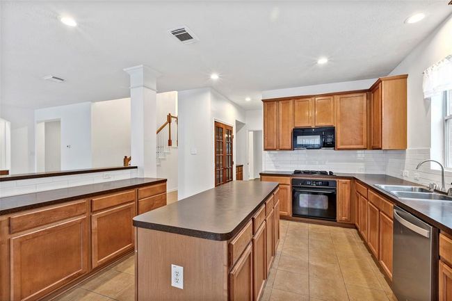 This kitchen features warm wooden cabinetry, a central island with dark countertops, and modern appliances including a stove, microwave, and dishwasher. It's a spacious, open layout with tile flooring, recessed lighting, and plenty of natural light from a window near the sink. | Image 6