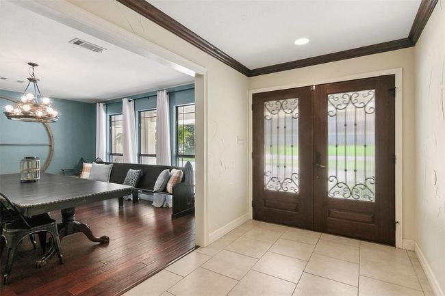 This inviting entryway features elegant double doors with decorative glass panels, opening into a spacious room with rich wood flooring. The adjacent dining area, accented with a modern chandelier, offers ample natural light and a cozy built-in bench. | Image 8