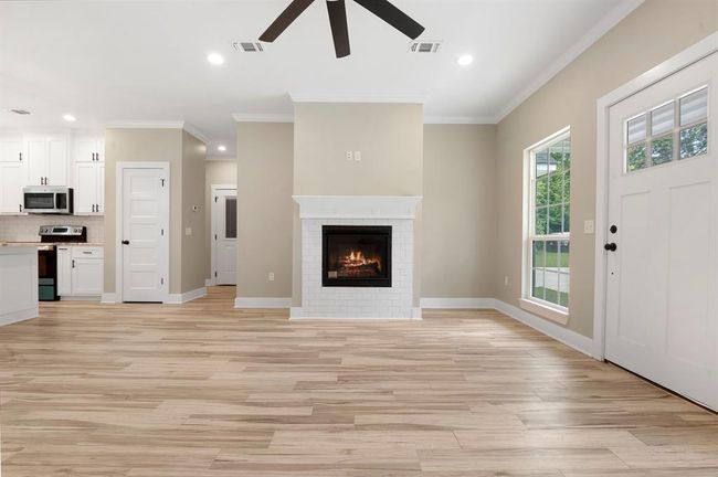 This bright, open-concept living space features a cozy fireplace, light wood flooring, and a ceiling fan. The adjacent kitchen has modern white cabinetry and stainless steel appliances, enhancing the home's contemporary appeal. Large windows and a front door with glass panels allow natural light to fill the room. | Image 4