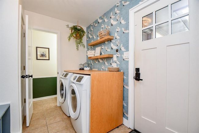 This photo showcases a bright and stylish laundry room featuring a modern washer and dryer set. The space is accentuated by a decorative wallpaper with a bird motif, open wooden shelving for storage, and a potted plant adding a touch of greenery. A door with glass panels provides natural light, enhancing the room's welcoming atmosphere. | Image 39