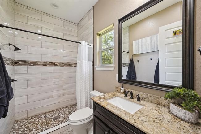 This bathroom features a modern walk-in shower with stylish tile work and a pebble floor. It includes a granite countertop, a large framed mirror, and a vanity with dark wood cabinetry. Natural light streams in through the window, enhancing the warm, inviting atmosphere. | Image 8