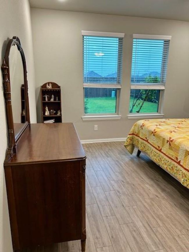 This cozy bedroom features wood-look tile flooring, two large windows offering plenty of natural light, and a view of the backyard. It includes a classic wooden dresser with a mirror and a bed adorned with a colorful quilt. | Image 14