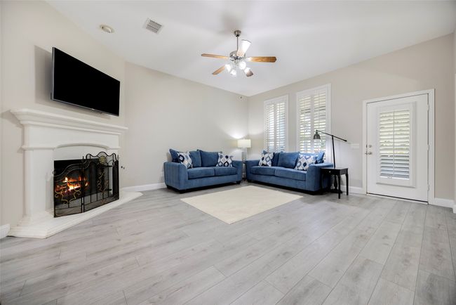 Living room featuring ceiling fan, a warm lit fireplace, light wood-type flooring, and view of the beautiful patio/yard. | Image 8