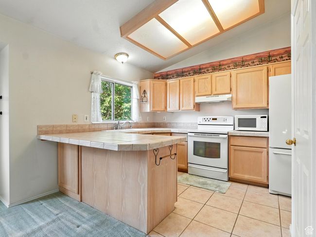 Kitchen with white appliances, under cabinet range hood, light brown cabinetry, lofted ceiling, and a peninsula | Image 4