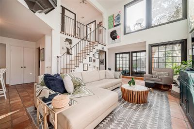 This is a bright and airy living room featuring high ceilings, large windows with ample natural light, Saltillo tile floors, a comfortable seating area, and a stylish staircase leading to an upper level. | Image 2