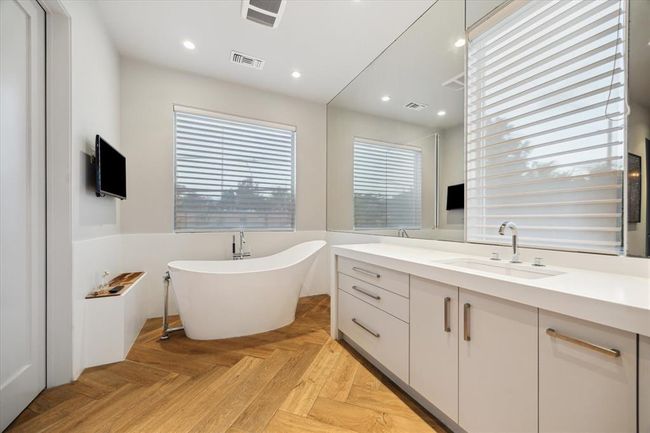 Sleek and serene, this side of the primary bath features a deep freestanding soaking tub with modern fixtures, a wall-mounted TV for added luxury, and expansive windows with soft-filtered natural light. A floating vanity with clean-lined cabinetry and contemporary hardware complements the warm herringbone wood-look flooring. A spa-like retreat designed for relaxation and style. | Image 17