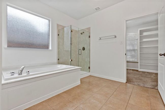 This photo showcases a spacious bathroom featuring a large soaking tub, a glass-enclosed shower, and a tile floor. The room is bright with a frosted window, and there's an open doorway leading to a carpeted area with built-in shelving. | Image 15