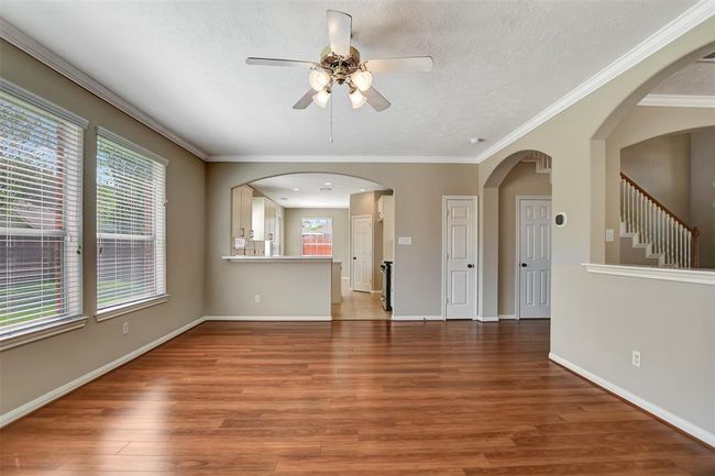 Bright and spacious living area with hardwood floors, large windows, and a ceiling fan. It features an open layout leading to the kitchen with a breakfast bar. Neutral colors and elegant crown molding enhance the welcoming atmosphere. | Image 16