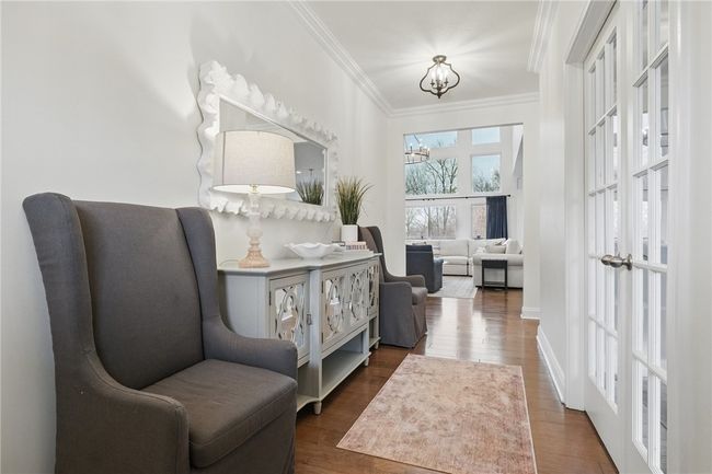 Bright and elegant entry hallway featuring hardwood floors and stylish lighting. French doors lead to a versatile room, with the space opening into a sunlit living area showcasing soaring ceilings and large windows with serene views. | Image 5