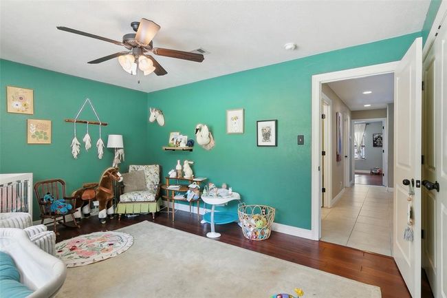 Bright and cheerful nursery with teal walls and hardwood floors, featuring playful decor and ample space for activities. The room includes a ceiling fan, plush toys, and a cozy seating area, with an open doorway leading to a hallway. | Image 32