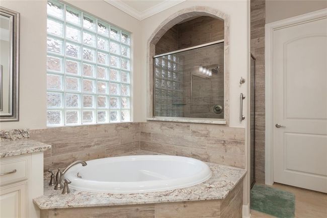 This bathroom features a luxurious soaking tub with stone tile surround, a large glass block window for natural light, and an adjacent walk-in shower with glass doors. The neutral tones and granite countertops add elegance and a spa-like atmosphere. | Image 31